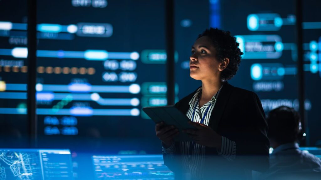 A woman in a darkly lit room surrounded by monitors and data panels.