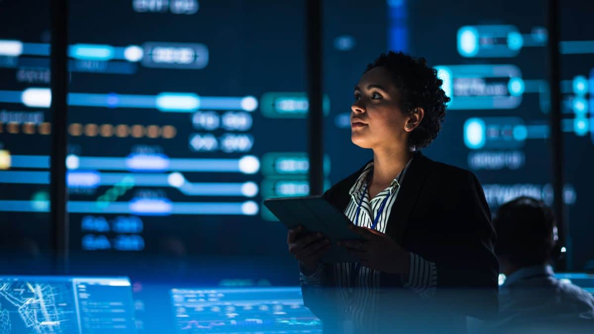 A woman in a darkly lit room surrounded by monitors and data panels.