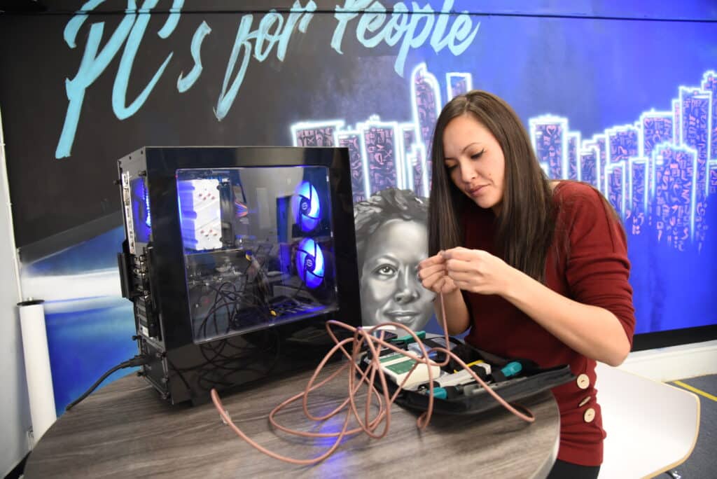 An ActivateWork learner working with wires and a motherboard in front of a wall painted with a PCs for People mural
