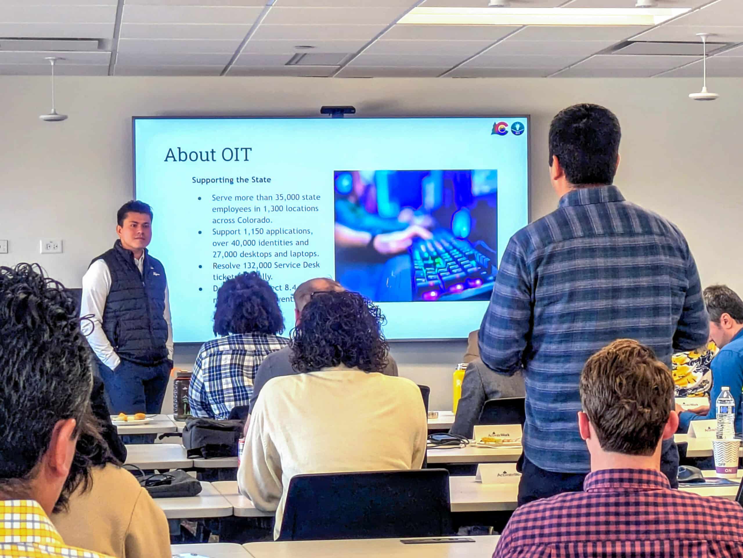 A man wearing a blue plaid shirt stands and addresses a question to a man in a black vest at the front of a classroom of adult learners. A slide on the presentation screen reads,