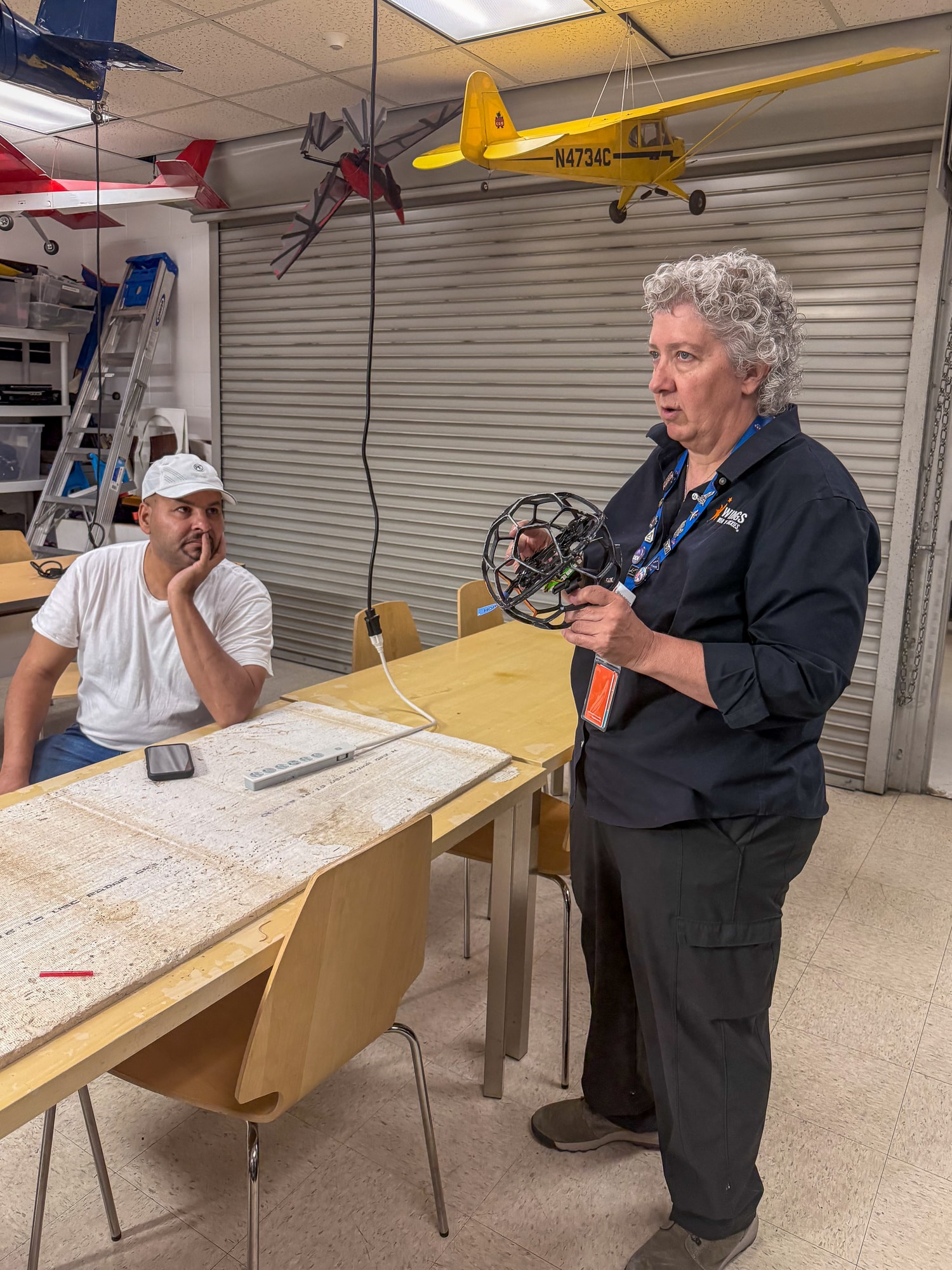 A woman wearing a Wings Over the Rockies uniform and a name tag lanyard is standing beside a long worktable, holding a geometric model and speaking to people outside the frame of the photo. Various airplane models hang from the ceiling.